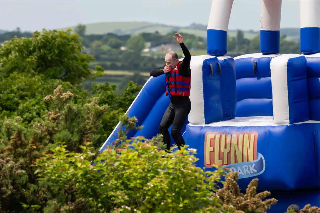 Man in life jacket jumps from a blue inflatable at Flynn Park, surrounded by lush greenery.