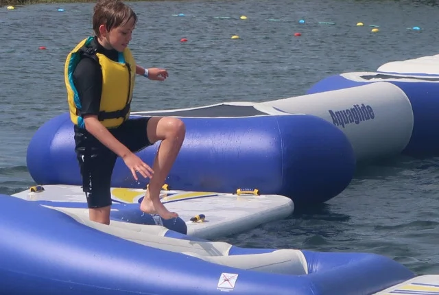 Boy in a yellow life jacket navigating an inflatable water course on a sunny day.