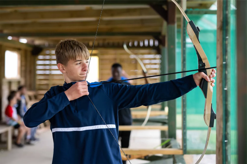 Teenager aiming a bow and arrow indoors at an archery range.