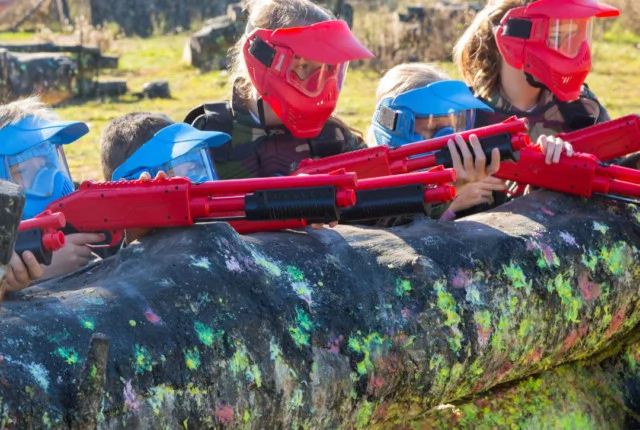 Children in colourful helmets play paintball, holding red paintball guns behind a splatter-painted obstacle.