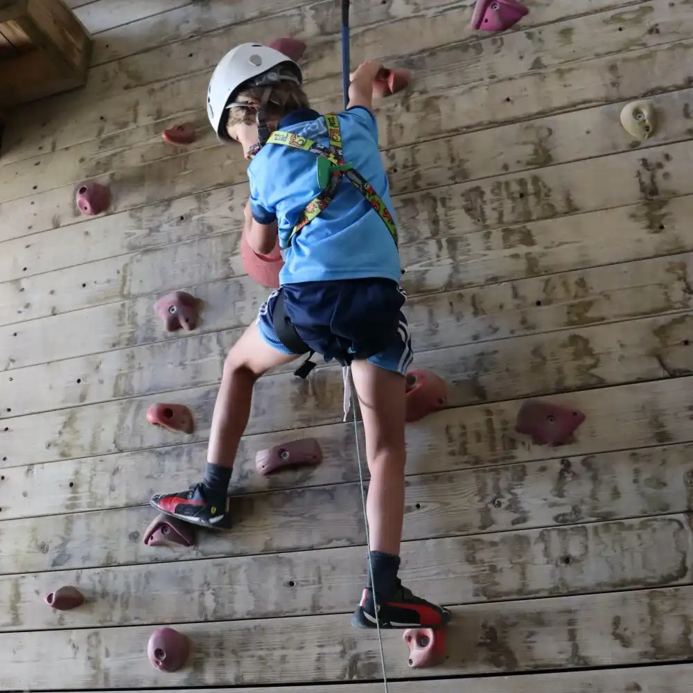 Child climbing a wooden wall with a safety harness and helmet.