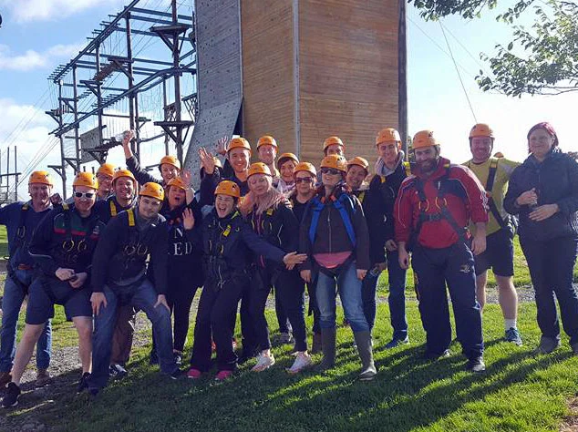Group of people in helmets and harnesses smiling outdoors near a high ropes course in the sunshine.