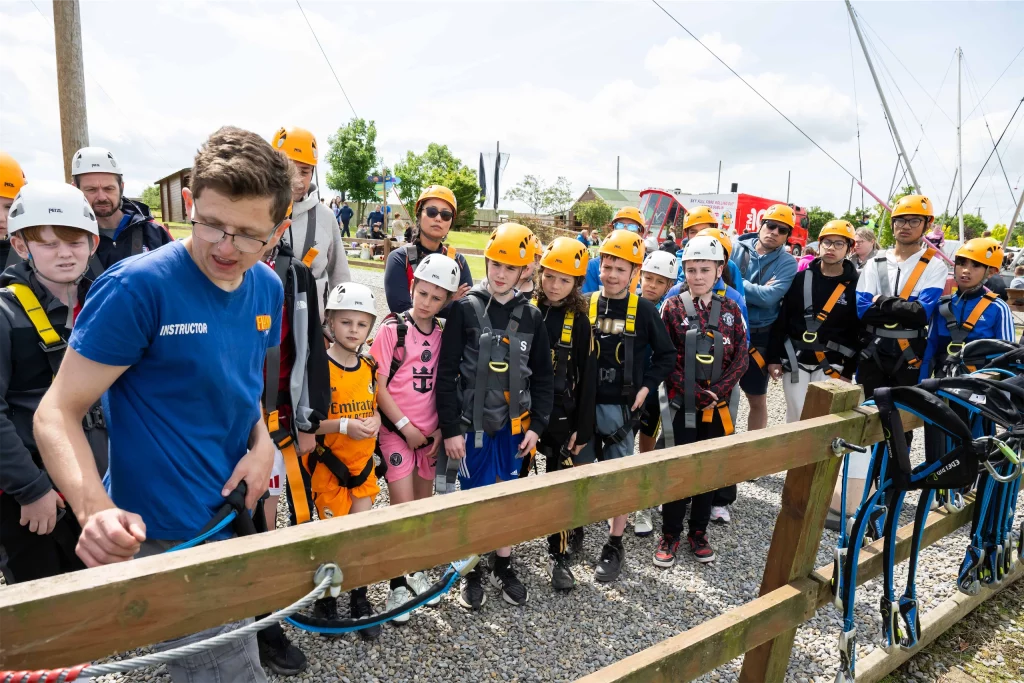 Group of children and a guide gather around for a zipline safety briefing, all wearing helmets and harnesses.