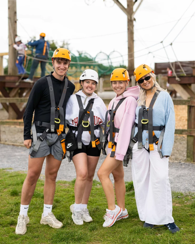 Four friends in helmets and harnesses ready for a high ropes course, smiling and standing on grass.