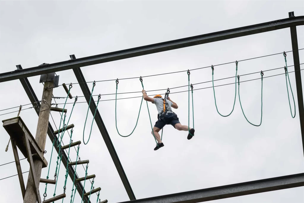 Person balancing on high ropes course under cloudy sky, wearing safety harness and helmet.