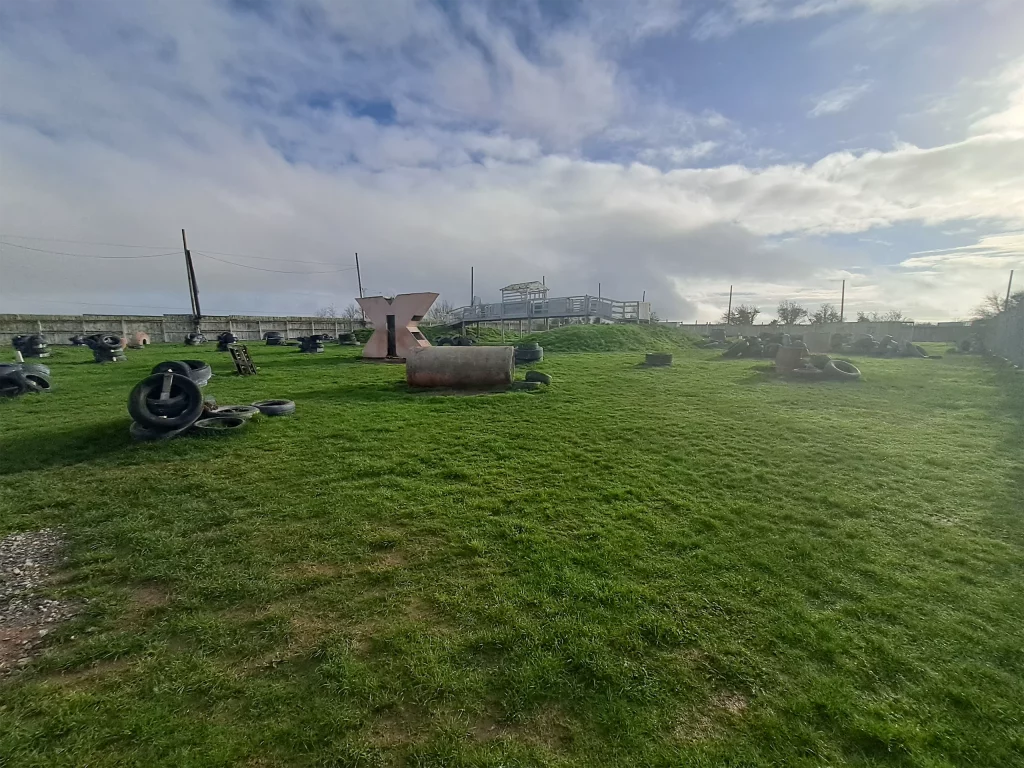 Grassy paintball field with tyre obstacles under a partly cloudy sky.