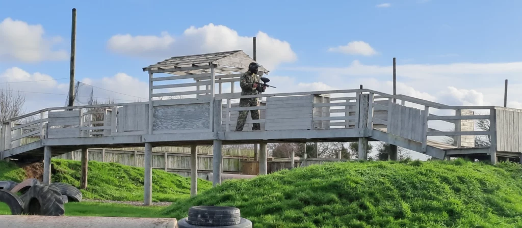Person in camouflage stands on a wooden platform with a paintball gun, surrounded by green grass and blue sky.
