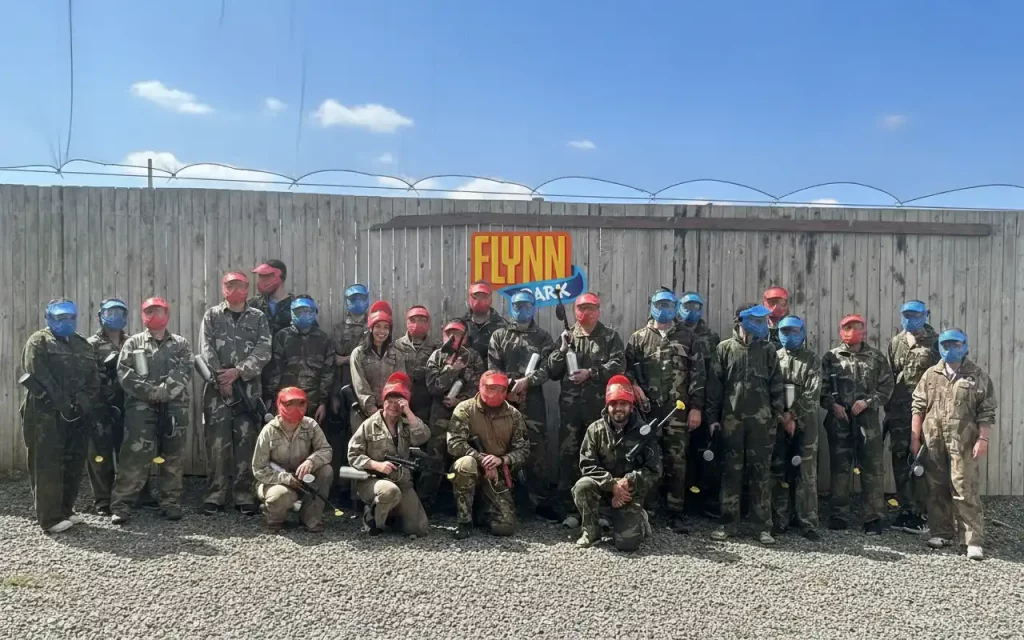 Group of paintball players in camouflage gear and helmets, standing by a wooden fence under a bright blue sky.