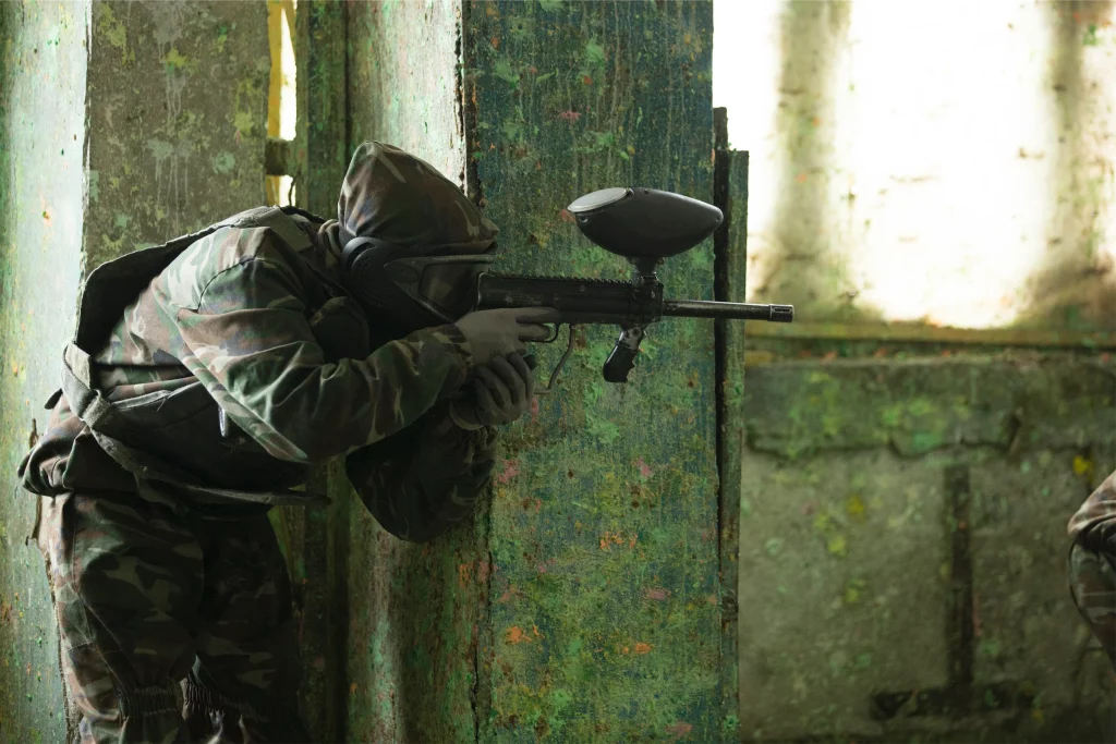 Person in camouflage gear aiming a paintball marker in an indoor arena with paint-splattered walls.