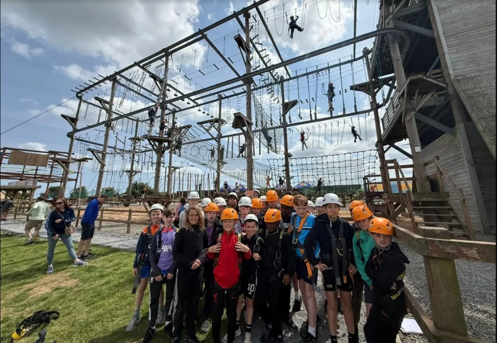 Group of kids in helmets and harnesses at an adventure park high ropes course, under a bright sky.