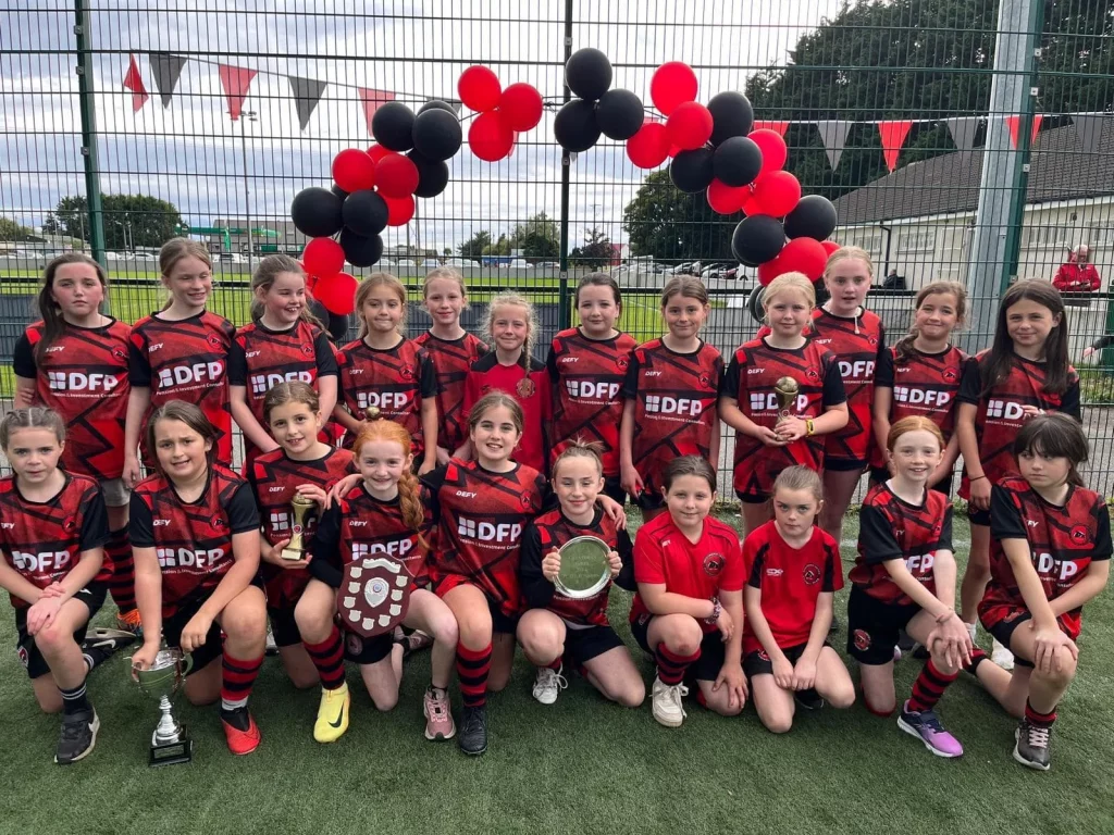 Girls' football team in red and black kits celebrating with trophies and shields under balloon arch on a sports field.