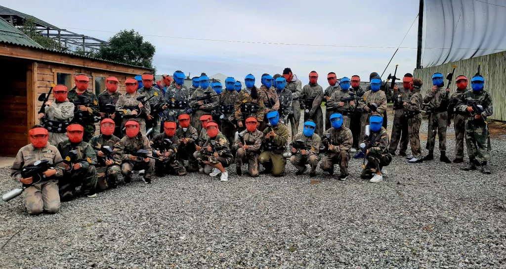 Group in camouflage gear with paintball guns, wearing red and blue masks, standing outdoors on gravel.