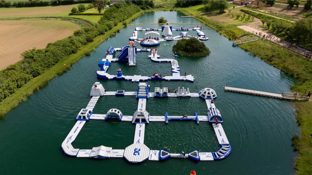 Floating inflatable water park with slides and obstacles on a lake, surrounded by lush green fields and trees.