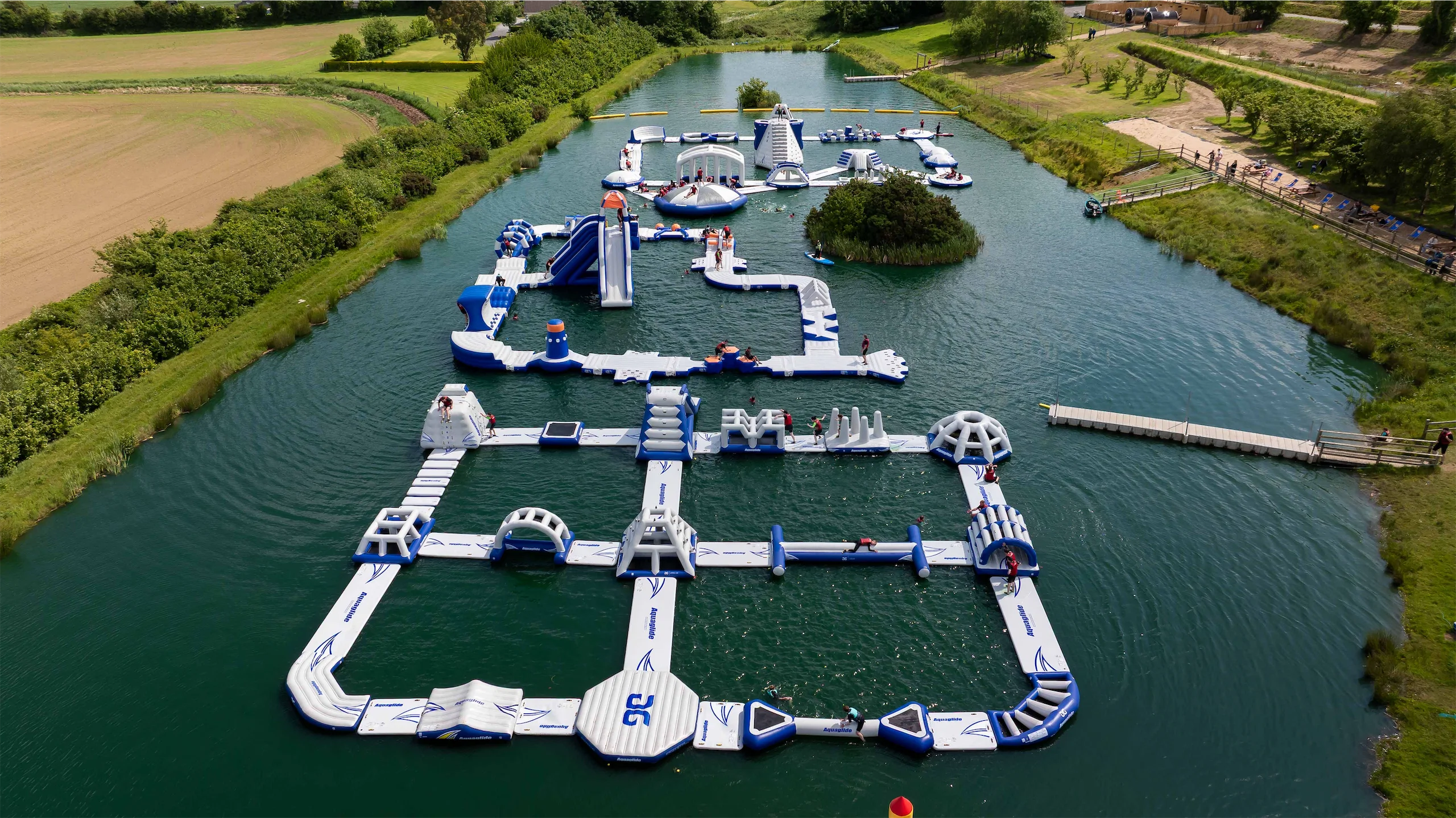 Floating inflatable water park with slides and obstacles on a lake, surrounded by lush green fields and trees.