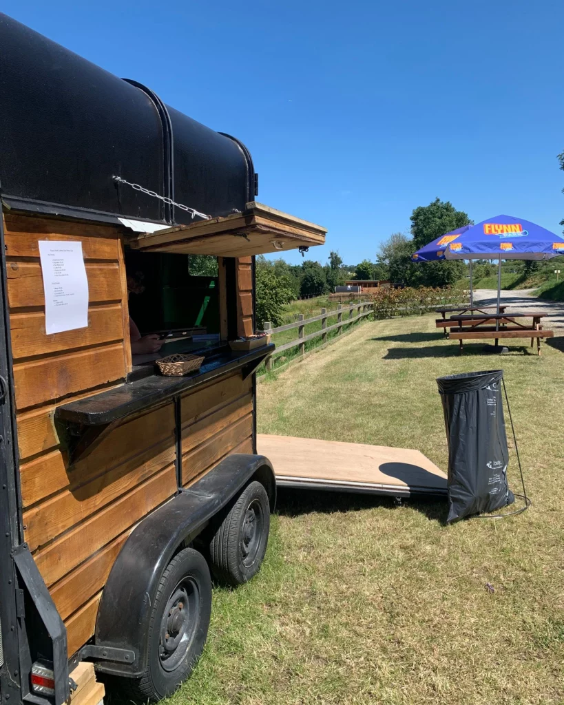 Food truck in a grassy field with picnic tables, parasol, and clear blue sky in the background.