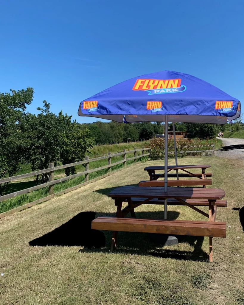 Wooden picnic tables with a blue parasol in a sunny park, surrounded by green fields and a wooden fence.