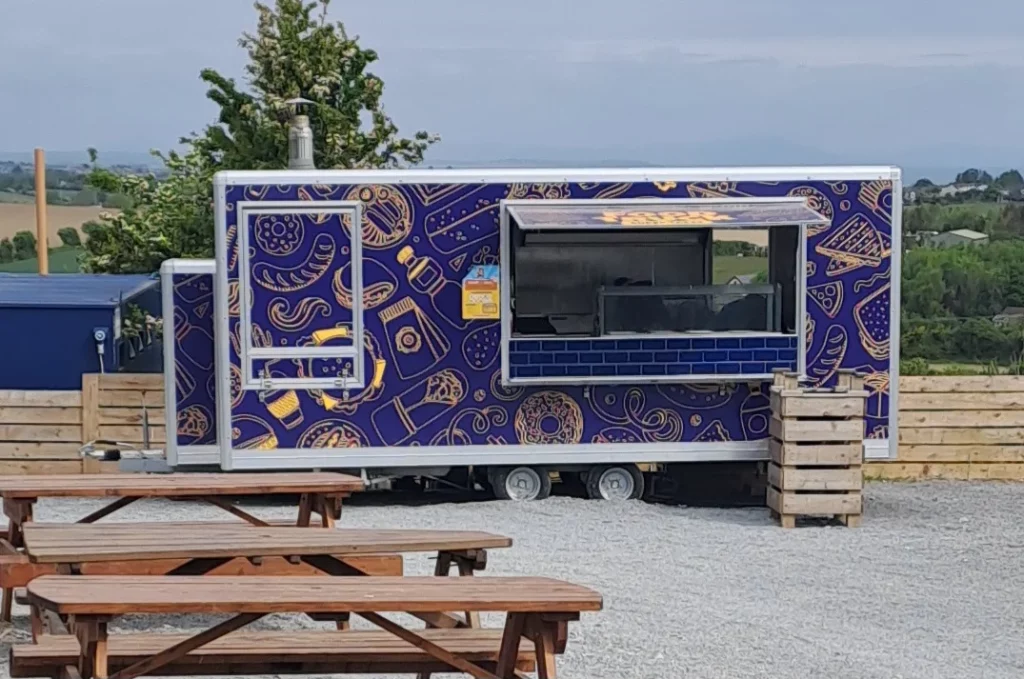 Colourful food truck with patterns, set against a rural landscape and wooden picnic tables in the foreground.