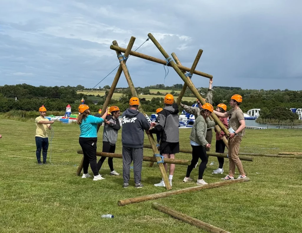 Group on grassy field building a wooden structure wearing orange helmets; teamwork in outdoor activity.
