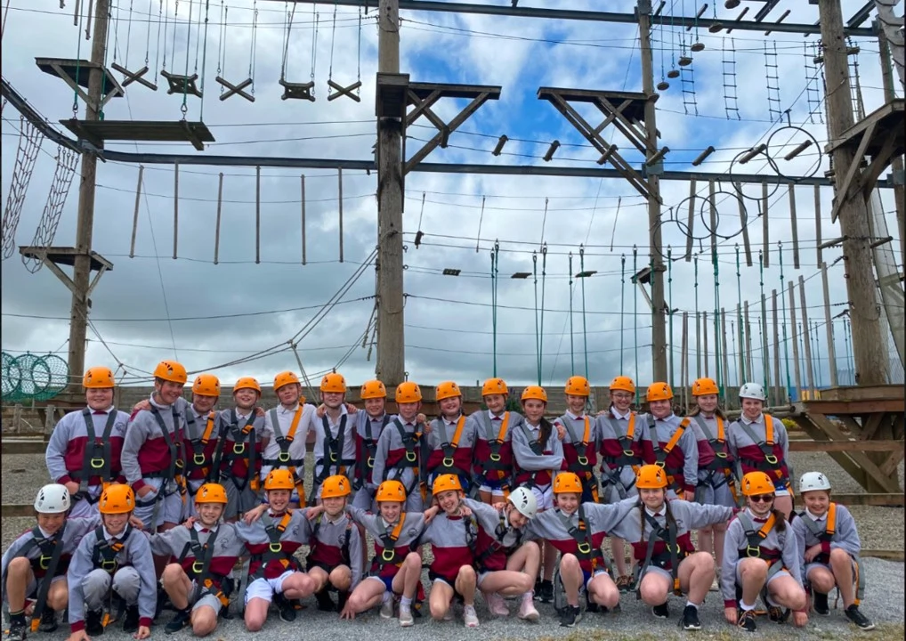 Group of teens wearing helmets and harnesses at an outdoor high ropes course, smiling under a cloudy sky.