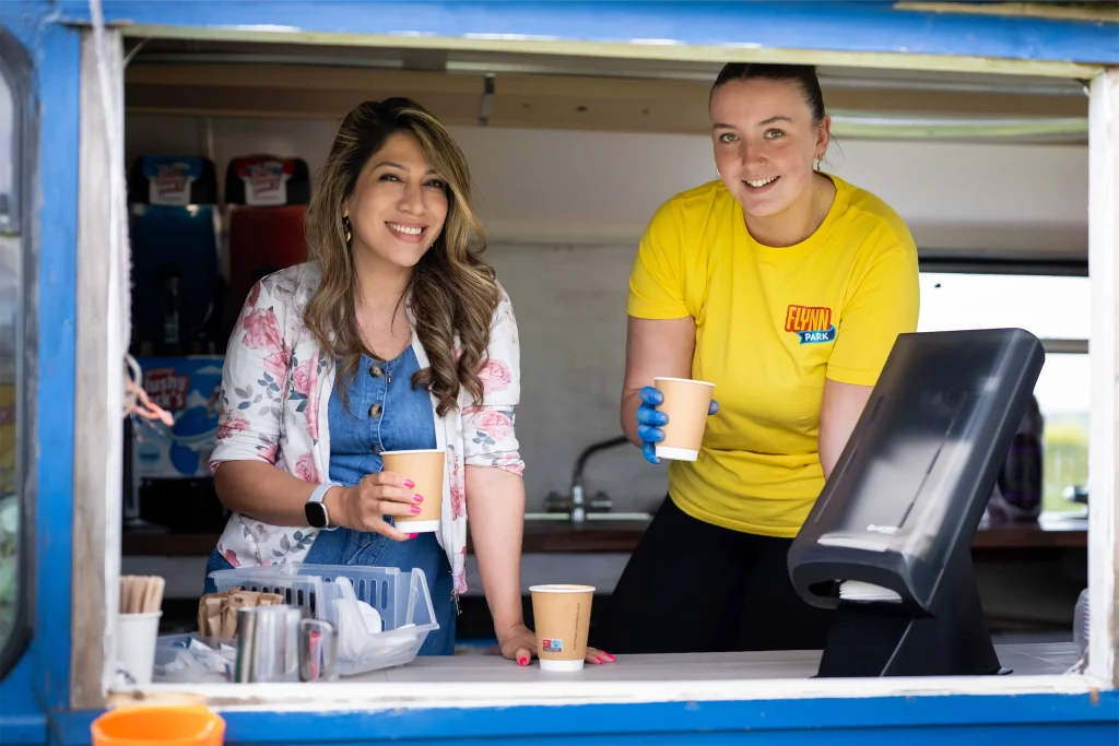 Two women smiling in a food truck, one wearing a floral jacket and the other in a yellow shirt with a coffee cup.