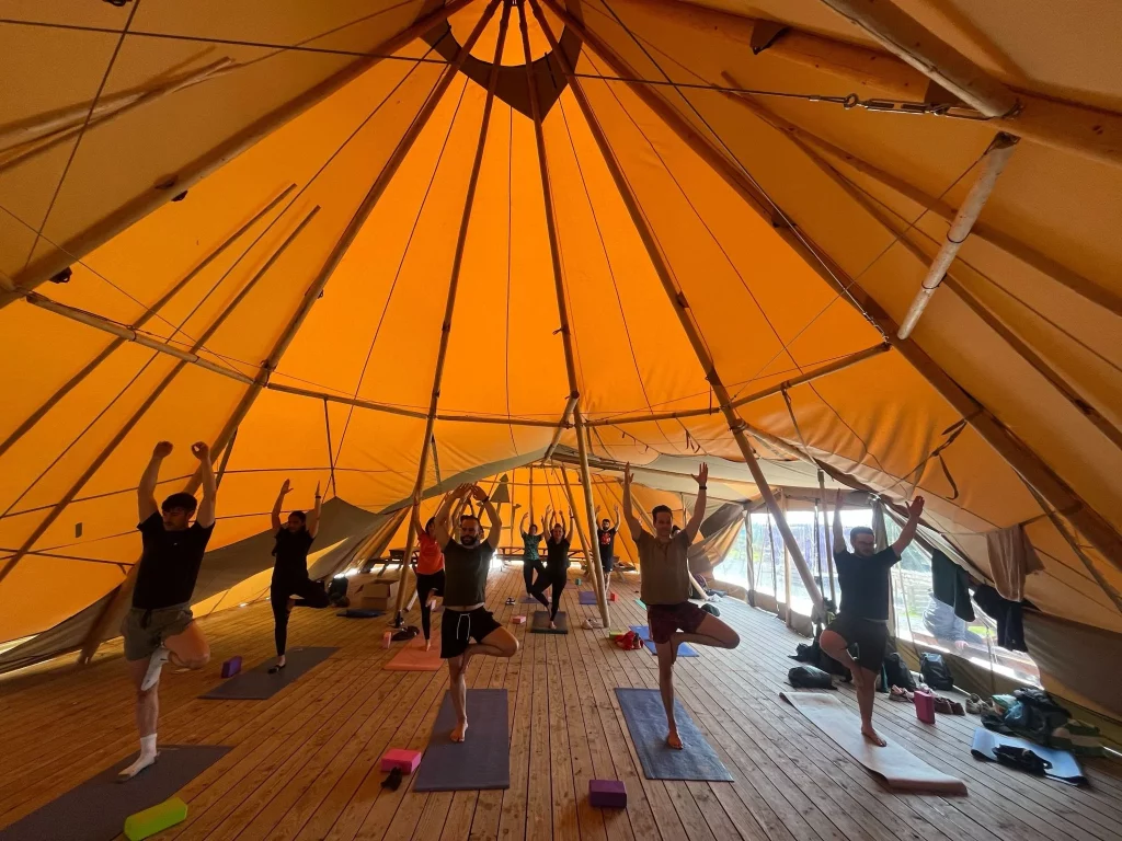 Yoga class under a large yellow tent, participants in tree pose on yoga mats.