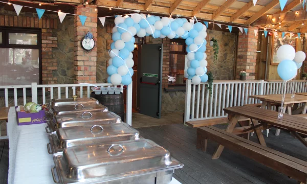 Buffet table set up on wooden deck with blue and white balloon arch, fairy lights, and picnic benches.