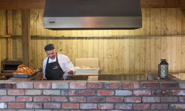 Chef in a rustic kitchen preparing food with a tray of fresh bread and a brick oven.